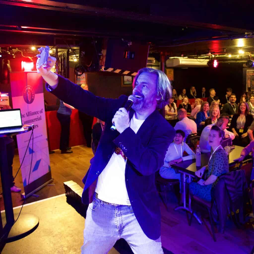 David McGuire onstage at a conference, taking a selfie with the audience while holding a microphone. The venue is a comedy club, with blue stage lighting. Suspiciously, he’s wearing the same blazer and T-shirt as in the first picture.
