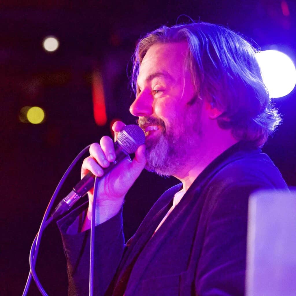 David McGuire, speaking into a microphone under red and blue lighting. He’s hosting a conference in a comedy club.