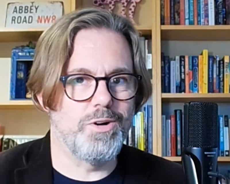Thumbnail for a short video about training, with a play button. David’s sitting behind a microphone, wearing serious glasses and that blazer again. On shelves behind him, there’s an assortment of children’s books, writing manuals, a TARDIS, and the Abbey Road sign.